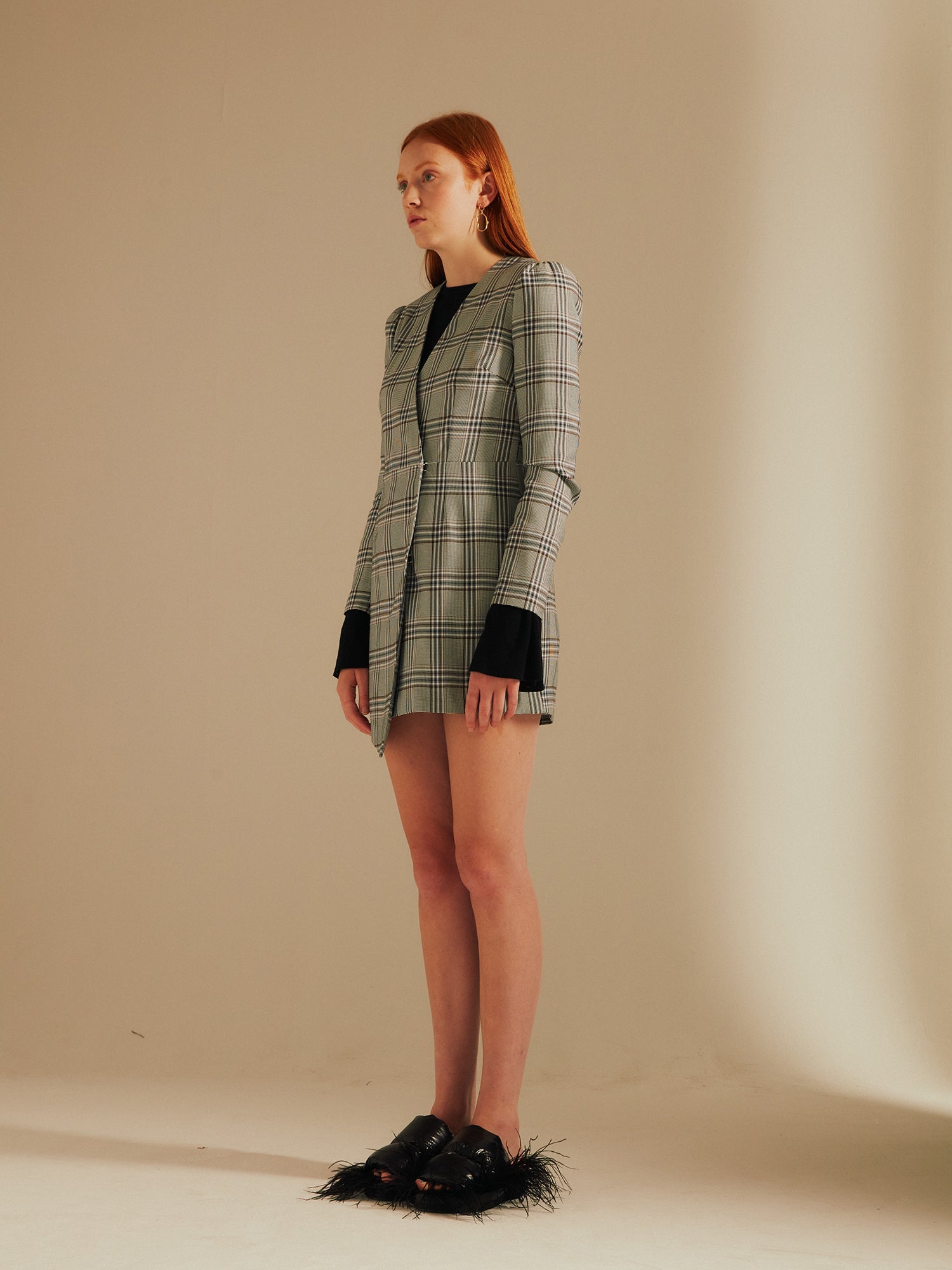 model faces away wearing the Amelia Heritage Asymmetric Blazer Dress with fluffy slippers in a studio